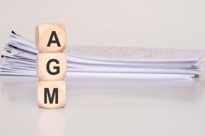 Three wood cubes stacked with letters agm placed in front of a stack of documents on a desk clean