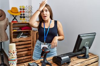 Młoda brunetka trzyma sztandar z otwartym tekstem w sklepie detalicznym zaskoczona ręką na głowie za pomyłkę, pamiętaj o błędzie. zapomniałem, zła koncepcja pamięci.
