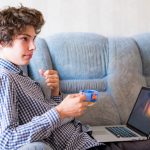 Młody uśmiechnięty nastolatek studiuje w domu z długie włosy używać laptopu obsiadaniem na kanapie