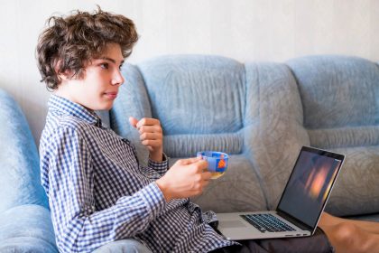 Młody uśmiechnięty nastolatek studiuje w domu z długie włosy używać laptopu obsiadaniem na kanapie