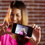 Uśmiechnięta azjatykcia kobieta bierze selfie z smartphone na ściana z cegieł