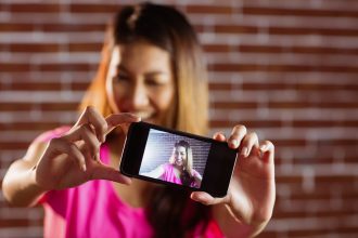 Uśmiechnięta azjatykcia kobieta bierze selfie z smartphone na ściana z cegieł