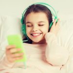 ludzie, dzieci, odpoczynek i koncepcja technologii - szczęśliwa uśmiechnięta dziewczyna leżąca na jawie ze smartfonem i słuchawkami w łóżku słuchająca muzyki w domu