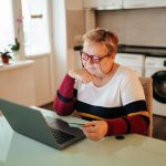 Starsza kobieta wyglądająca na nadwagę siedzi w domu i używa laptopa, trzymając w ręku kartę kredytową, robiąc zakupy online za pośrednictwem handlu elektronicznego