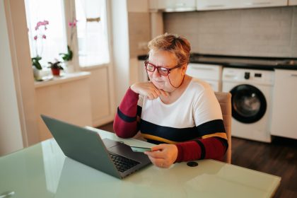 Starsza kobieta wyglądająca na nadwagę siedzi w domu i używa laptopa, trzymając w ręku kartę kredytową, robiąc zakupy online za pośrednictwem handlu elektronicznego