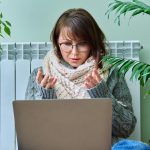 Kobieta w średnim wieku w ciepłych ubraniach ogrzewa się w pobliżu grzejnika używając laptopa do rozmowy wideo konferencja czat sezon jesień zima technologia w wolnym czasie praca styl życia 40-latków koncepcja