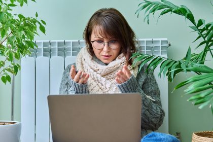 Kobieta w średnim wieku w ciepłych ubraniach ogrzewa się w pobliżu grzejnika używając laptopa do rozmowy wideo konferencja czat sezon jesień zima technologia w wolnym czasie praca styl życia 40-latków koncepcja