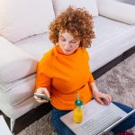 Freelancer kobiet ładny czerwony rude włosy, czytanie wiadomości tekstowych, siedząc na podłodze z otwartym komputerem w nowoczesnym domu, młoda kobieta kreatywna praca na laptopie, mając sok pomarańczowy