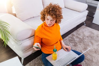 Freelancer kobiet ładny czerwony rude włosy, czytanie wiadomości tekstowych, siedząc na podłodze z otwartym komputerem w nowoczesnym domu, młoda kobieta kreatywna praca na laptopie, mając sok pomarańczowy