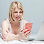 Blondynka w domowych ubraniach leży na łóżku ze smartfonem w dłoni i laptopem. Praca zdalna podczas pandemii koronawirusa.