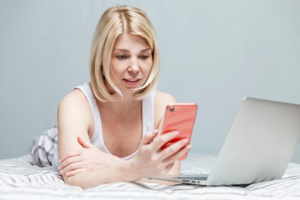 Blondynka w domowych ubraniach leży na łóżku ze smartfonem w dłoni i laptopem. Praca zdalna podczas pandemii koronawirusa.