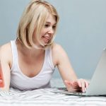Dorosła kobieta rasy białej w piżamie jest podekscytowana, że znalazła dobre wiadomości w Internecie za pomocą laptopa, trzymając w dłoni smartfon. Miejsce na tekst.