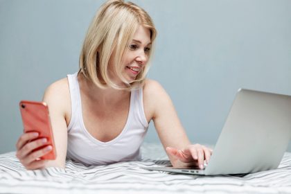 Dorosła kobieta rasy białej w piżamie jest podekscytowana, że znalazła dobre wiadomości w Internecie za pomocą laptopa, trzymając w dłoni smartfon. Miejsce na tekst.