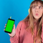 Piękna młoda kobieta słucha muzyki przez słuchawki i okulary przeciwsłoneczne, chromowany ekran telefonu komórkowego