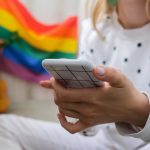 Młoda milenijna hippie siedząca na balkonie z telefonem komórkowym lgbtq tęcza flaga
