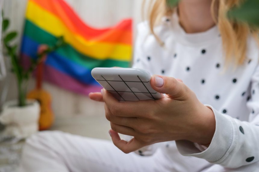 Młoda milenijna hippie siedząca na balkonie z telefonem komórkowym lgbtq tęcza flaga